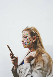 Frau mit Aufklebern im Gesicht, die ein Smartphone vor einem weißen Hintergrund benutzt - SVCF00245