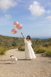 Verspieltes Mädchen im weißen Kleid hält Luftballons mit Hund auf dem Weg - MEGF00316