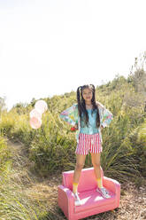 Smiling girl standing with arms akimbo on pink sofa in meadow - MEGF00324