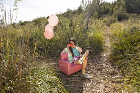 Mädchen, das durch eine Spielzeugkamera fotografiert und auf einem rosa Sessel in einem Feld sitzt - MEGF00328