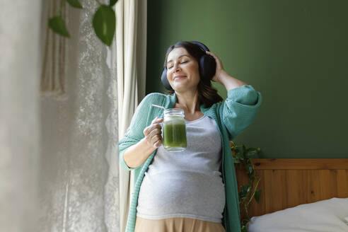 Lächelnde schwangere Frau hört Musik über drahtlose Kopfhörer und hält einen grünen Smoothie in der Hand - TYF00499