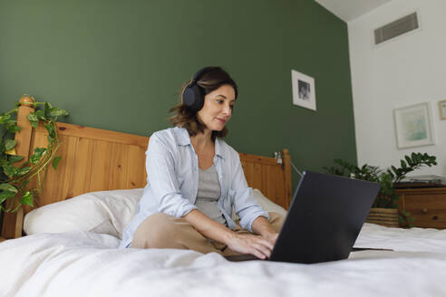 Pregnant businesswoman wearing wireless headphones using laptop on bed in bedroom - TYF00501