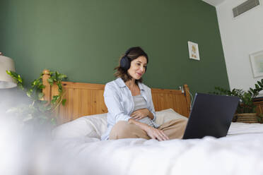 Lächelnde schwangere Geschäftsfrau mit drahtlosen Kopfhörern und Laptop auf dem Bett im Schlafzimmer - TYF00502