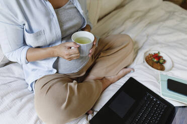 Pregnant businesswoman holding tea cup sitting with laptop on bed - TYF00503
