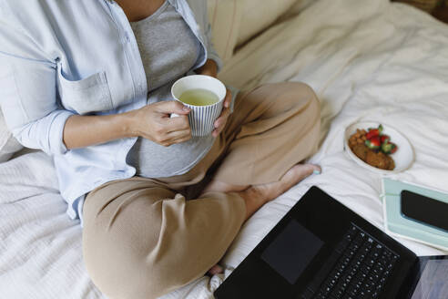 Schwangere Geschäftsfrau mit Teetasse und Laptop auf dem Bett sitzend - TYF00503