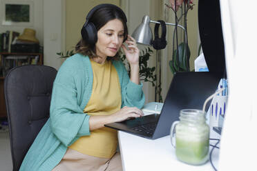 Pregnant businesswoman wearing wireless headphones using laptop at home office - TYF00509