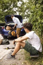 Side view of teenage boy sitting on skateboard looking at father loading luggage in car trunk - MASF33448