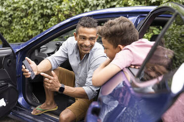 Excited father showing mobile phone to son leaning on car door - MASF33457