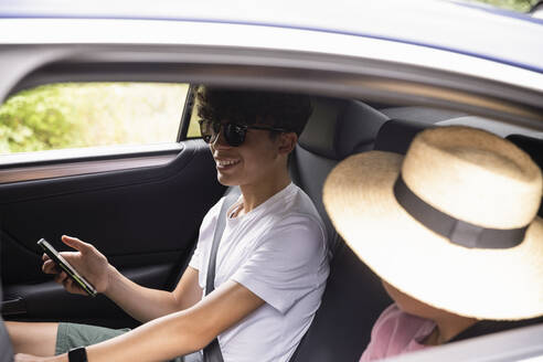 Lächelnder Jugendlicher mit Smartphone und Sonnenbrille, der neben seinem Bruder im Auto sitzt - MASF33459