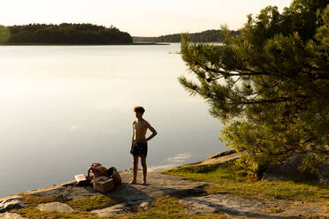 Shirtless teenage boy with hand on hip standing at lakeshore during vacation - MASF33486