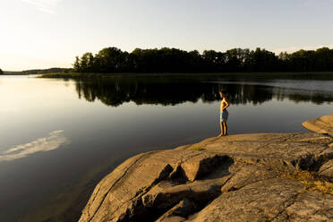 Junge ohne Hemd schaut auf den See, während er im Urlaub auf einem Felsen steht - MASF33487