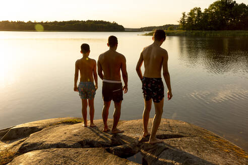 Rückansicht eines Vaters mit Söhnen auf einem Felsen bei Sonnenuntergang im Urlaub - MASF33490