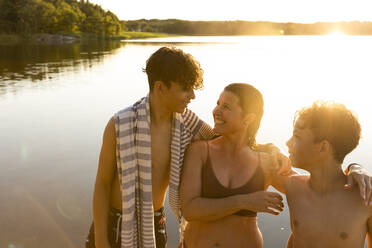 Glückliche Söhne im Gespräch mit ihrer Mutter, während sie bei Sonnenuntergang am See stehen - MASF33500