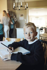 Porträt eines lächelnden Jungen, der mit einem Arbeitsbuch auf einem Tisch zu Hause sitzt - MASF33511