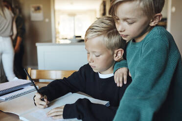 Junge mit Hand auf der Schulter seines Bruders, der zu Hause Hausaufgaben in einem Buch macht - MASF33513