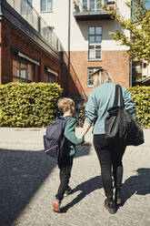 Rear view of mother walking with son towards school building on sunny day - MASF33538