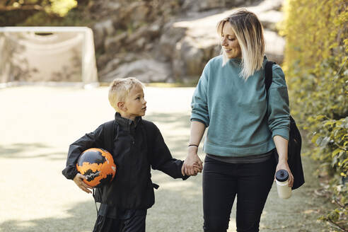 Mutter hält die Hand ihres Sohnes, der einen Fußball trägt, während er auf dem Boden läuft - MASF33543