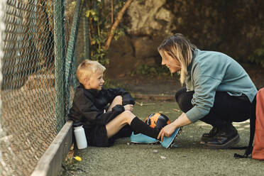 Happy woman tying shoelace of son sitting by fence in ground - MASF33544