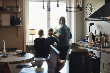 Rear view of mother with sons looking outside from kitchen window - MASF33545