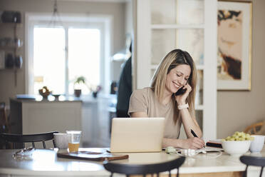 Happy freelancer taking down notes while talking on smart phone at home - MASF33548