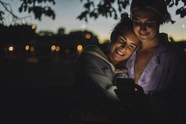 Lächelnde Frau umarmt nicht-binären Freund mit Handy in der Nacht - MASF33618