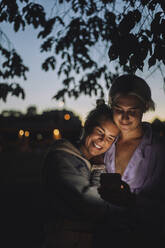Lächelnde nicht-binäre Person, die nachts das Mobiltelefon einer Freundin benutzt - MASF33619