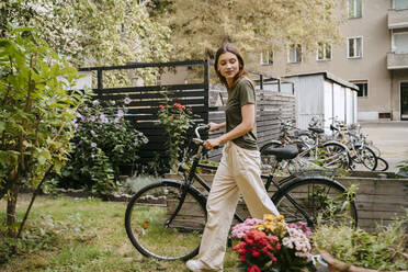 Seitenansicht einer jungen Frau, die mit dem Fahrrad fährt und Blumen im Garten betrachtet - MASF33622