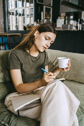 Young female entrepreneur writing in book holding coffee cup while sitting on sofa in studio - MASF33638