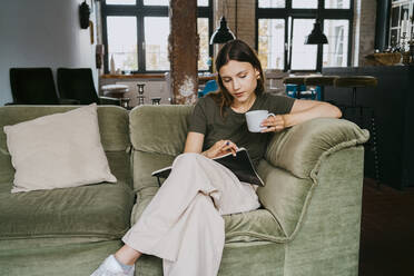 Junger Unternehmer liest ein Buch und trinkt einen Kaffee, während er auf dem Sofa im Studio sitzt - MASF33639