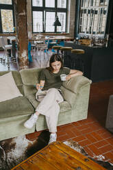 Entrepreneur sitting on sofa with legs crossed while reading book and having coffee in studio - MASF33640