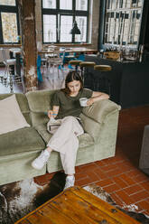 Unternehmer sitzt mit gekreuzten Beinen auf dem Sofa, liest ein Buch und trinkt Kaffee im Studio - MASF33640