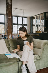 Female using smart phone and having coffee while sitting with legs crossed on sofa in studio - MASF33642