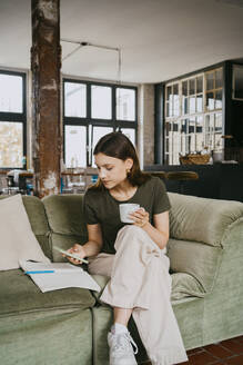 Frau, die ein Smartphone benutzt und einen Kaffee trinkt, während sie mit gekreuzten Beinen auf dem Sofa im Studio sitzt - MASF33642