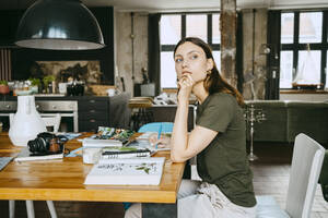Weiblicher Food-Stylist mit Hand am Kinn, der wegschaut, während er am Tisch im Studio sitzt - MASF33654