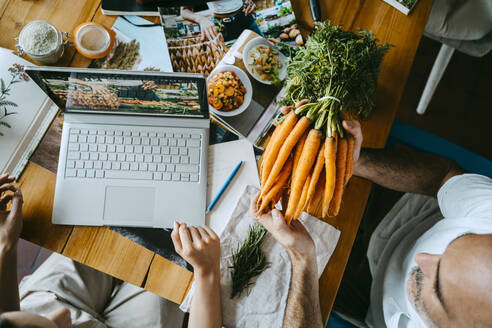 Ein männlicher Kollege hält ein Bündel Karotten in der Hand, während ein Unternehmer einen Laptop auf dem Tisch hat - MASF33666
