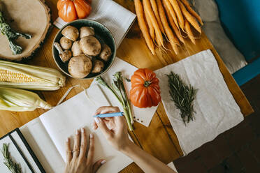 Blick von oben auf einen Foodstylisten, der inmitten von Gemüse auf einem Tisch Tagebuch schreibt - MASF33674