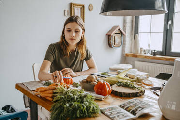 Junge Unternehmerin mit Karotten in der Hand am Tisch sitzend im Studio - MASF33675