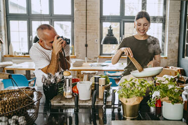 Glückliche Köchin beim Kochen mit einem männlichen Kollegen, der sie im Studio fotografiert - MASF33680