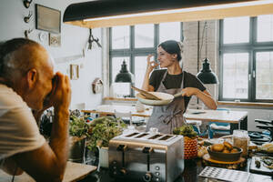Männlicher Fotograf fotografiert eine Köchin, die in einer Studioküche eine Pfanne hält und gestikuliert - MASF33689