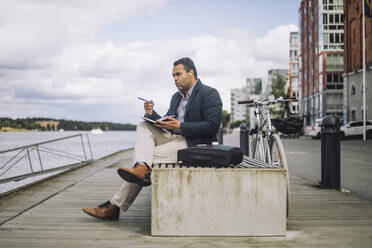 Full length of male freelancer sitting with diary on bench by bicycle at promenade - MASF33696
