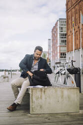 Full length of male entrepreneur searching in backpack while sitting on bench at promenade - MASF33697