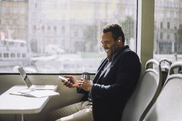 Cheerful male entrepreneur with mobile phone and insulated drink container sitting at table in ferry - MASF33708