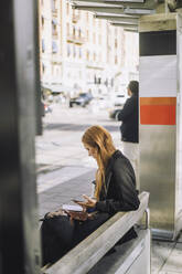 Side view of female freelancer sitting with smart phone at bus stop - MASF33709