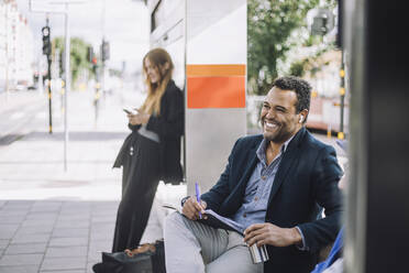 Glücklicher männlicher Freiberufler mit kabellosen In-Ear-Kopfhörern, der mit einem Tagebuch gegen eine Geschäftsfrau an der Bushaltestelle sitzt - MASF33710