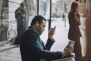 Seitenansicht eines männlichen Berufstätigen, der eine Voicemail sendet, während er an einer Bushaltestelle gegenüber einer Geschäftsfrau sitzt, die mit einem Mobiltelefon spricht - MASF33714