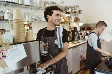Lächelnder Besitzer mit Schürze, der wegschaut, während er in der Nähe der Kaffeemaschine im Café steht - MASF33729