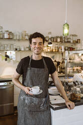 Lächelnder männlicher Besitzer mit Kaffeetasse in einem Café stehend - MASF33757