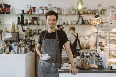 Lächelnder männlicher Besitzer mit Schürze, der eine Kaffeetasse in einem Café hält - MASF33758