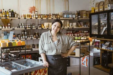 Portrait of female owner with hand on hip standing by ice cream freezer in store - MASF33785