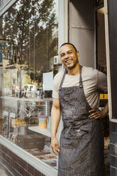 Portrait of smiling male deli owner with hand on hip by doorway - MASF33800